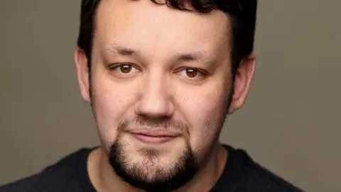 Wolf Marloh Joe Staton looks at the camera. He has brown eyes, dark brown hair, a brown beard and is wearing a black t-shirt.