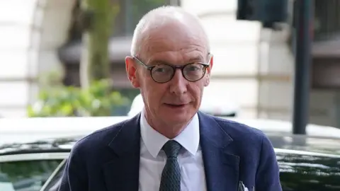 PA Pat McFadden, wearing a pair of glasses with brown rims, pictured with a car and a street background behind him