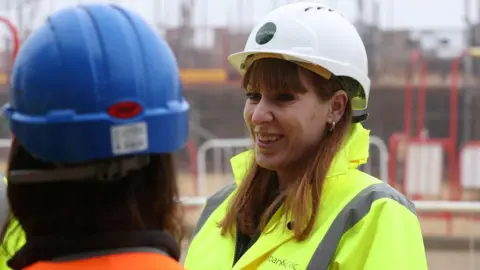 PA Media Angela Rayner wears a white hard hat and yellow hi-vis jacket. She is looking to the left of the camera and smiling at person in the foreground. 