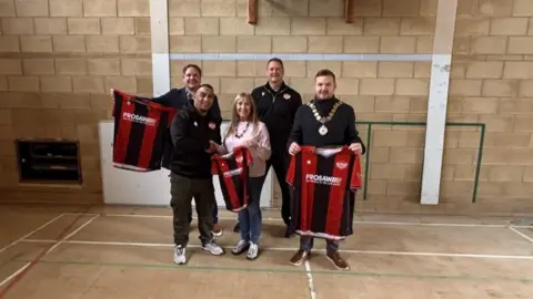 Four men, all dressed in black tops, and a woman, in a pink jumper, stand in a sports hall holding up three red and black football shirts. 