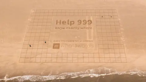 What3Words A large piece of sand art showing a grid of more than 100 boxes measuring 3m square. In a large central box there is lettering reading Help 999 know exactly where - Every 3m square has an address' and the What3Words logo.