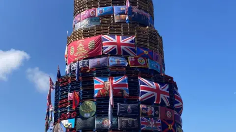 A large number of flags including union flags and a flag depicting an image of King Charles placed on a bonfire in Derry's Bogside.