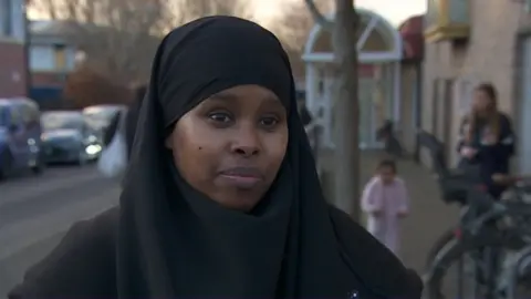 A Muslim woman is looking at the camera. She is wearing a black hijab. She is stood in front of Easton Leisure centre entrance. 