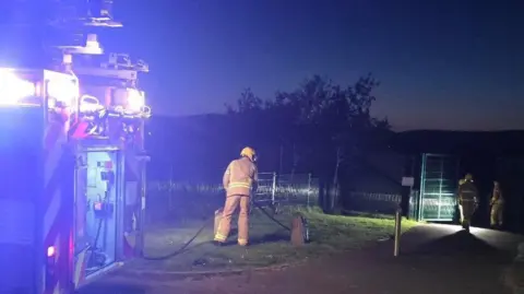 Mark H Durkan A number of firefighters stand near to a fire engine with its lights flashing.
A hose can be seen being brought towards the gates of the play park.