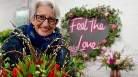 A woman smiles as she stands in a flower show behind a basket of red tulips and in front of a wall decoration shaped like a heart with pink fluorescent letters reading "Feel the love" and a border of green leaves and pink roses. She has short white hair and wears black-framed glasses, a blue coat and matching scarf.