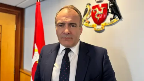 Alfred Cannan is wearing a dark blue suit, white shirt and blue tie with small red spots. He is clean shaven with dark receding hair. He is standing in an office with a large Manx flag to the left and an Isle of Man crest on the wall to the right.