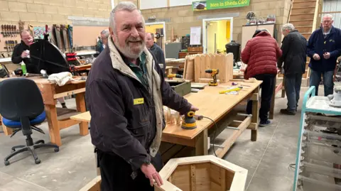 An older man stood around a wooden planter - which he is making - and smiling as he rests his hand on a nearby drill on a wooden bench. Other men can be seen in the workshop, working on their own projects.