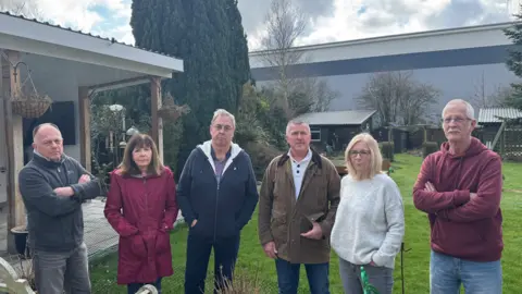 George King/BBC Four men and two women standing in a row in a back garden. The large warehouse can be seen in the background.