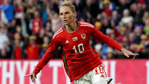 PA Media Jess Fishlock holds both arms out to celebrate scoring a goal for Wales. She wears Wales' football kit and a blurred background shows a stand full of people watching the game. 
