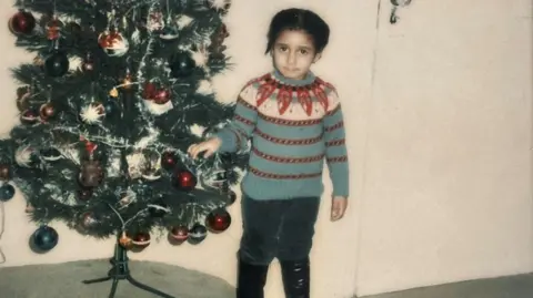 Metropolitan Police A polaroid image of a Andrea Bernard standing next to a decorated Christmas tree. She is wearing a patterned blue, white, and red knit jumper, dark trousers, and black boots.