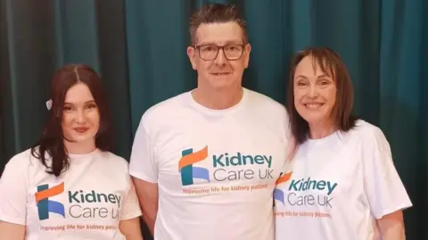 A young woman standing beside a man and another woman. The two women have brown hair and the man has short brown hair. They are all wearing white t-shirts with "kidney care UK" written on them.