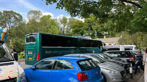 A large blue coach parked in a busy car park in Bilbury. There are lots of cars lining the road, and an ice cream truck parked to the left of the image.