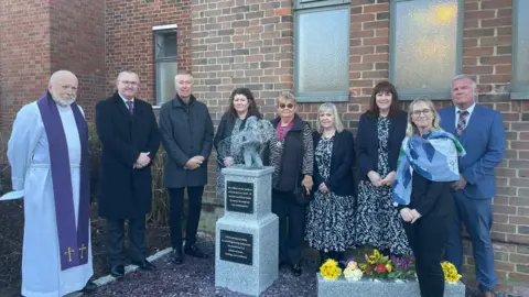 Colchester City Council Nine people, including a vicar, stand around the Covid-19 memorial. They are looking at the camera and smiling. The memorial consists of two rectangular marble plinths, one on top of the other. It is topped with the figure of a kneeling woman holding a dandelion. Behind the gathering, the side of a brick building can be seen.