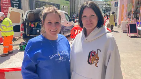 Two women stand side by side in front of the road closure. It is a sunny day and shops can be seen lining the street, with a small pavement area outside of the cordoned-off roadworks. Emily Marsland is wearing a blue jumper and has brown hair which is tied back. Charlotte Miles is wearing a beige hoodie and has short brown hair which she is wearing down.