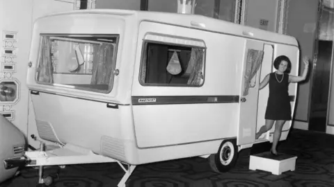 Daily Mirror/Mirrorpix via Getty Images A woman stands on the step of a 1969 Eccles Amethyst caravan waving at the camera. She is in a 1960s style dress with a large necklace chain around her neck.