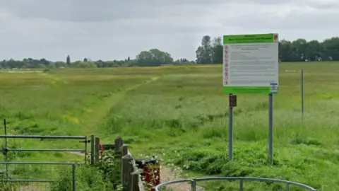 Google A wide open green field under a cloudy sky, with a narrow path cutting through the grass. In the foreground, there is a metal gate and fence leading into the meadow area. A large sign on two posts reads “Bury Field Common and Meadow” and includes rules and guidance for visitors. Trees line the horizon in the distance, framing the rural landscape.