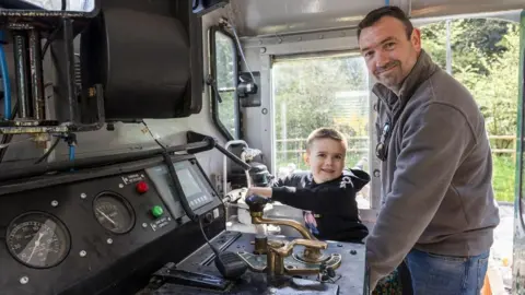 Cillian Gallagher and his father are in the locomotive playing with the buttons. They are both smiling at the camera.