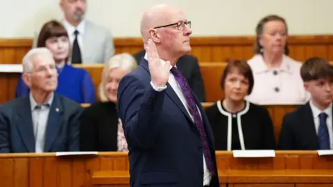 John Swinney, who is bald with glasses, stands side on with his right hand raised, with audience members of wooden benches looking on 