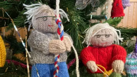 Knitted figures of Mr and Mrs Johnstone on a Christmas tree. He is wearing a grey sweater and blue trousers and holding a bell ringing rope. She is wearing a red jumper and knitting. 