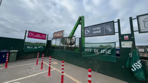 A household recycling Centre with bays for 'small electricals, 'wood and timber', and scrap metal'. In front are parking spaces and bright orange reflective bollards. The sky behind is overcast.