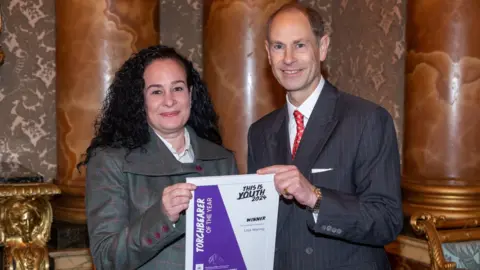 A lady with black curly hair standing next to the Duke of Edinburgh. She is wearing a light grey suit with purple lines and buttons.  The Duke of Edinburgh is wearing a dark grey pinstripe suit. They are both smiling at the camera and hold a certificate between them that is white and purple. 