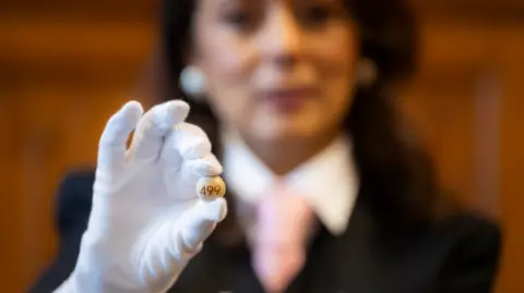 House of Commons A photo with a hand in a white glove holding a small ballot ball between finger and thumb is in the foreground. Behind and out of focus, is Nusrat Ghani, wearing her black hair loose around her shoulders, with a black jacket over a white shirt and a pink tie. 