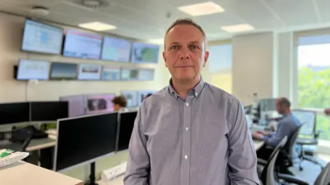 BBC A picture of Paul Aked stood in front of a number of computers and screens. Paul is wearing a grey shirt and he has short hair.