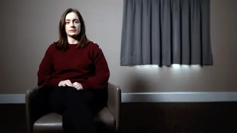 BBC business coach Rebecca Amin sits in a leather chair. He is wearing a maroon sweater with buttons on the shoulders. Behind him is a window with curtains closed.