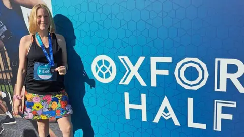 PA Media Toni Bull in black running vest and Mister Men shorts standing beside a blue 'Oxford Half Marathon' hoarding 