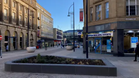 A flowerbed surrounded by a low granite wall in a city centre pedestrianised street.