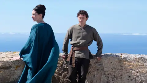 Tom Holland and Anne Hathaway in a film still from the film The Odyssey. Tom Holland is leaning against a wall with a scenic greek sea view behind him. He is wearing a dark olive green shirt and dark trousers. Anne hathaway is looking to the left and wearing a flowing flowing blue dress
