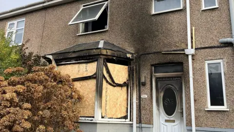 A brown, pebble-dashed, semi-detached house blackened by  smoke. The white front door is covered in smoke damage and the front bay window is boarded up.