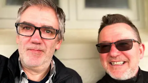 Stuart and Carl pose for a selfie. They both smile and are wearing glasses. 