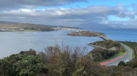 A grey road and bridge over a blue sea with some brown and green trees in the foreground, as white, fluffy clouds linger in the sky. 