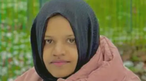 A woman in her 20s wearing a black hijab and pink puffy coat. There is grass behind her although the background is blurred. She has a slightly smiling but fairly neutral expression as she looks at the camera.