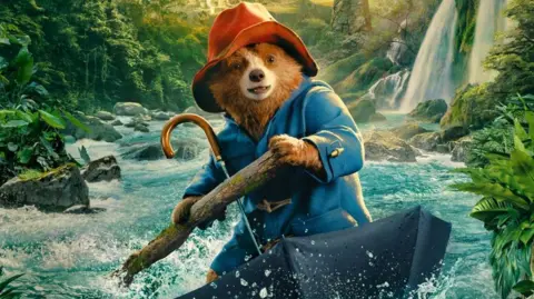 Paddington riding in a river in a umbrella holding a stick