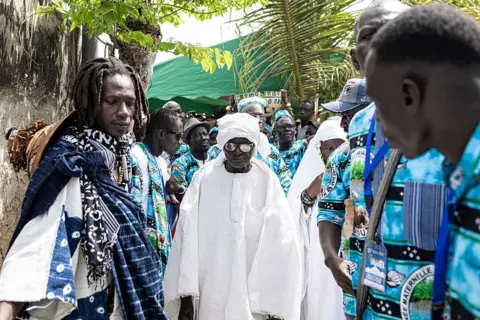 PATRICK MEINHARDT / AFP /GETTY IMAGES A man dressed in a white boubou walks past men dressed in matching blue clothing.