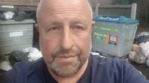 Darren Wilmot Headshot of a white man in a dark blue t-shirt infront of piles of rubbish and bins which have not been emptied.