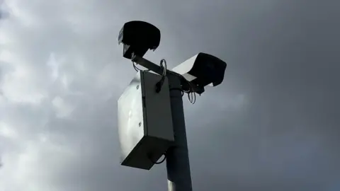 Henry Godfrey-Evans/BBC Two grey cameras at the top of a grey metal pole, against the backdrop of a moody grey sky.