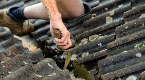 Getty Images A corrugated tiled roof is being repaired by a man whose knee and arm can be seen. He is holding a trowel which is scraping one of the tiles.