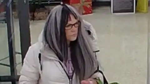 Rachael Cole wearing a white and black wig and glasses as she walks around a supermarket.