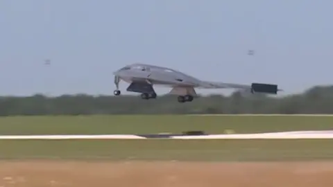 A grey war plane as it lands on a runway in a green field