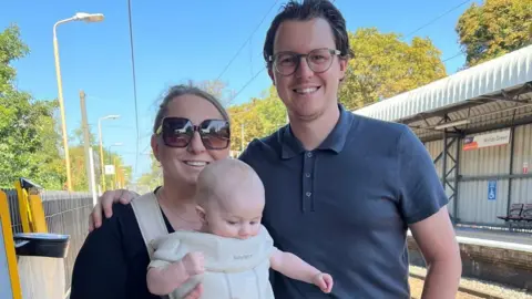 Laura in a black dress holding George in a cream baby carrier on her front with Daniel with his arm around her wearing shorts and a blue top, standing on a train platform