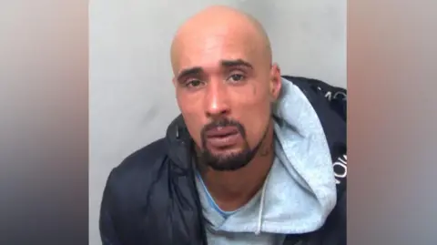 Essex Police A custody photo of Ashley Warren. He is wearing a grey hoodie with a black jacket over the top. He appears worried or concerned, facing the camera.