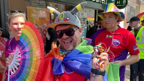 People dressed in rainbow colours, smiling on a hot summer's day. A man in the foreground is smiling, wearing black sunglasses. There are shops behind him.