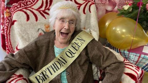 Gladys sitting in a chair wearing a 'Birthday Queen' sash and a tiara
