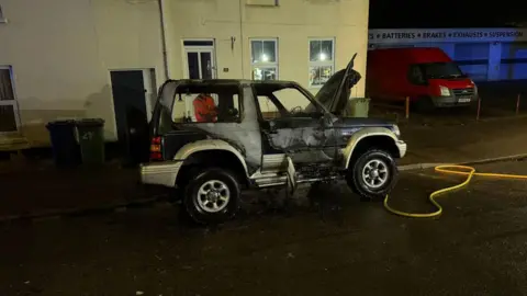 Wisbech Community Fire & Rescue Station The car after the flames were extinguished. The side of the car appears tarnished and the bonnet is open. There is a yellow fire hose on the ground in front of the car and a person in a hi-visibility orange jacket behind it. 