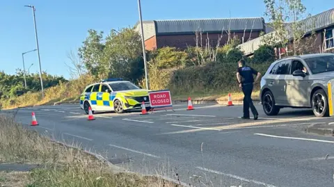 The image shows a road closure on a wide suburban road, with a marked police vehicle parked across part of the carriageway, traffic cones forming a barrier, and a red sign reading "ROAD CLOSED" in the middle of the road. On the right, a grey SUV has stopped near the closure while a police officer stands beside it.