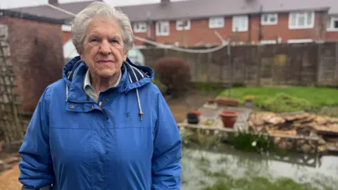 An elderly woman in a blue coat, stood to the left of her garden which is partially submerged underwater by the rain. She is stood in front of a brown fence and a row of houses.
