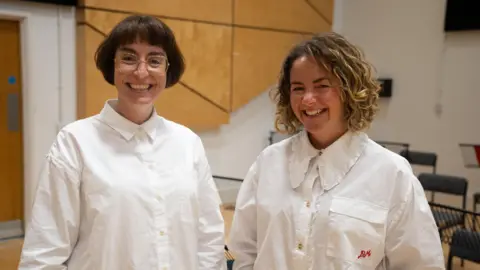 Adele Thomas and Sarah Crabtree stand side by side wearing white button-up shirts. Adele has short dark hair and glasses, and Sarah has curly blonde hair.